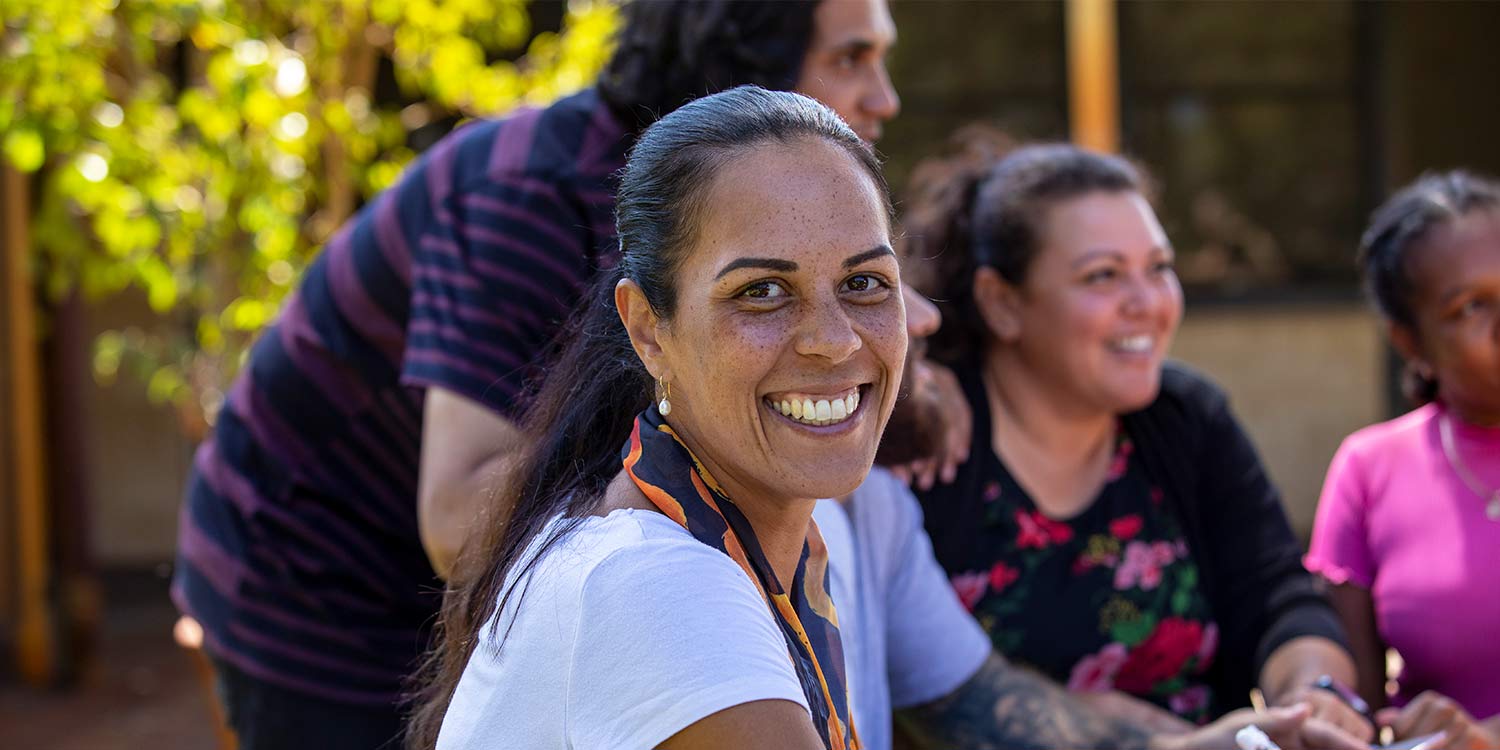 Close-up of aboriginal students and their tutor sitting outdoors in Australia. One of the female students is looking at the camera and smiling.