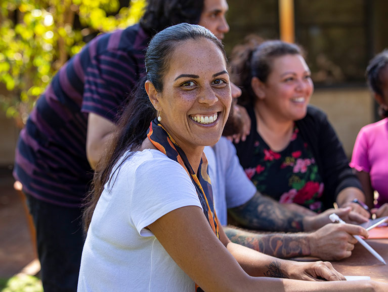 Close-up of aboriginal students and their tutor sitting outdoors in Australia. One of the female students is looking at the camera and smiling.